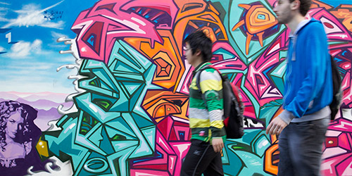 Two people walking past a wall decorated with vibrant and abstract graffiti. 