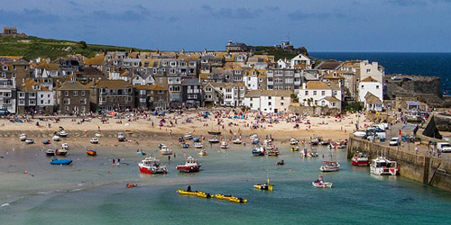 A panorama of St Ives bay in Cornwall. There are many people on the beach and many boats in the sea.