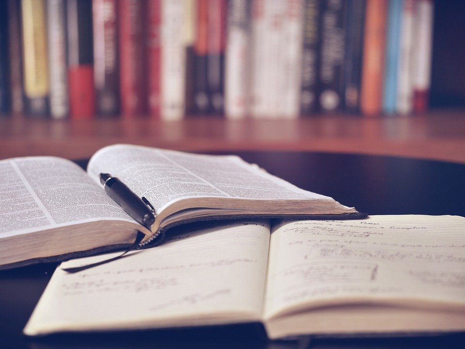 A desk with an open book, a notebook with writing in it and a pen. A bookshelf in the background.